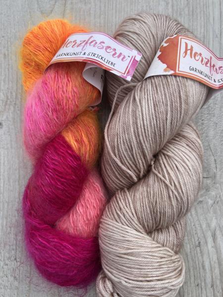Zwei Stränge handgefärbte Wolle: links Herzfasern No. 50 „Petrasfarben 2.0“ in leuchtendem Pink, Orange und Gelb mit Mohair-Struktur, rechts Herzfasern No. 11 „Dolomiten“ in hellem Beige auf glatter Merino-Basis. Fotografiert auf hellem Holzuntergrund.
